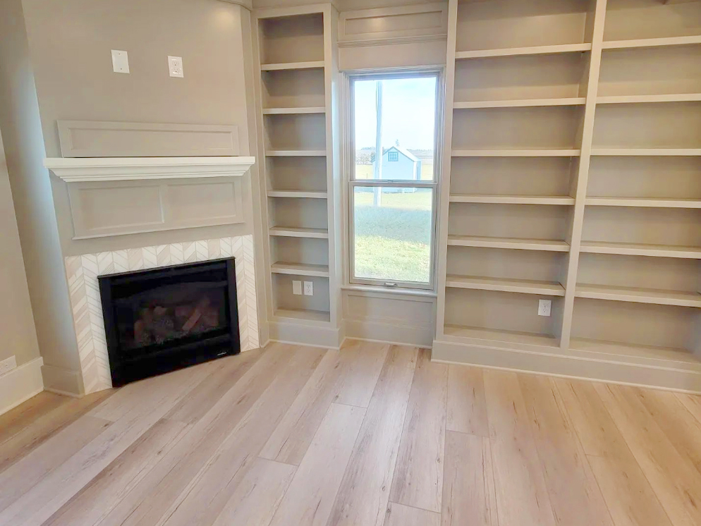 A living room features built-in shelving units surrounding a central window. Below the window is an electrical outlet. To the left is a fireplace with a mantle and a patterned tile surround. The room has light-colored wood flooring.