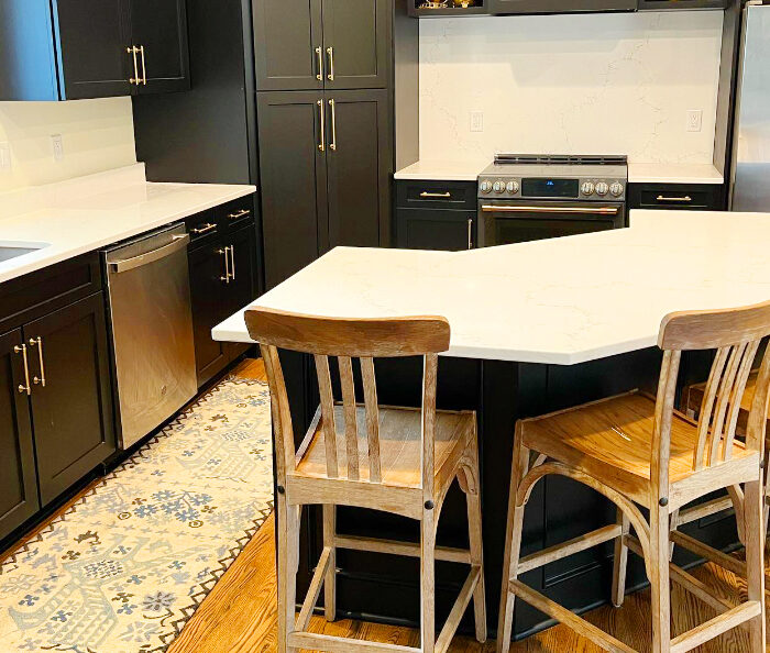 A modern kitchen with black cabinets, white countertops, and stainless steel appliances. A central island with a white countertop and wooden bar stools. A patterned rug is seen on the wooden floor near the sink and dishwasher.