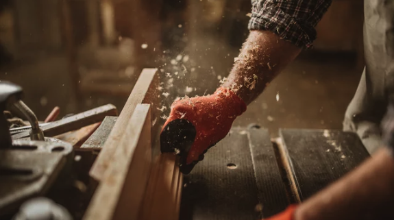 A person wearing red gloves uses a woodworking plane to smooth a piece of wood, causing wood shavings to fly. The scene is detailed and appears to take place in a well-equipped woodshop with various carpentry tools in the background.