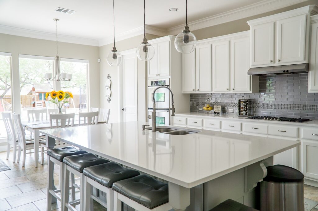 A modern kitchen with white cabinets, a large island with bar stools, stainless steel appliances, and pendant lighting. A dining area with a table and chairs is visible on the left, featuring a vase of sunflowers. The backsplash is gray subway tile.