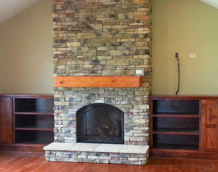 A stone fireplace extends upward to a vaulted ceiling with a wooden mantel. The stonework is composed of irregularly shaped stones in various shades of gray and brown. Flanking the fireplace are built-in wooden cabinets with shelves.