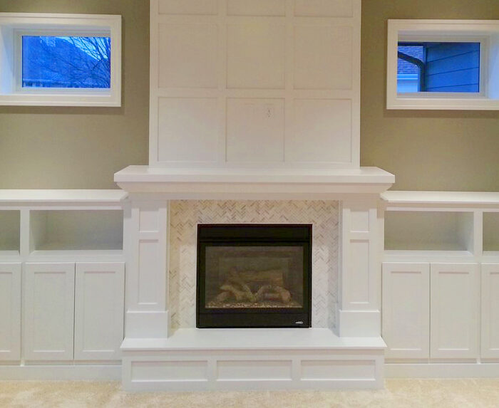 A modern living room features a white built-in fireplace with herringbone tile detail, flanked by symmetrical white cabinets and shelves. Above the fireplace, a large paneled wall extends up to the ceiling. Two small windows with blue-tinted evening light.