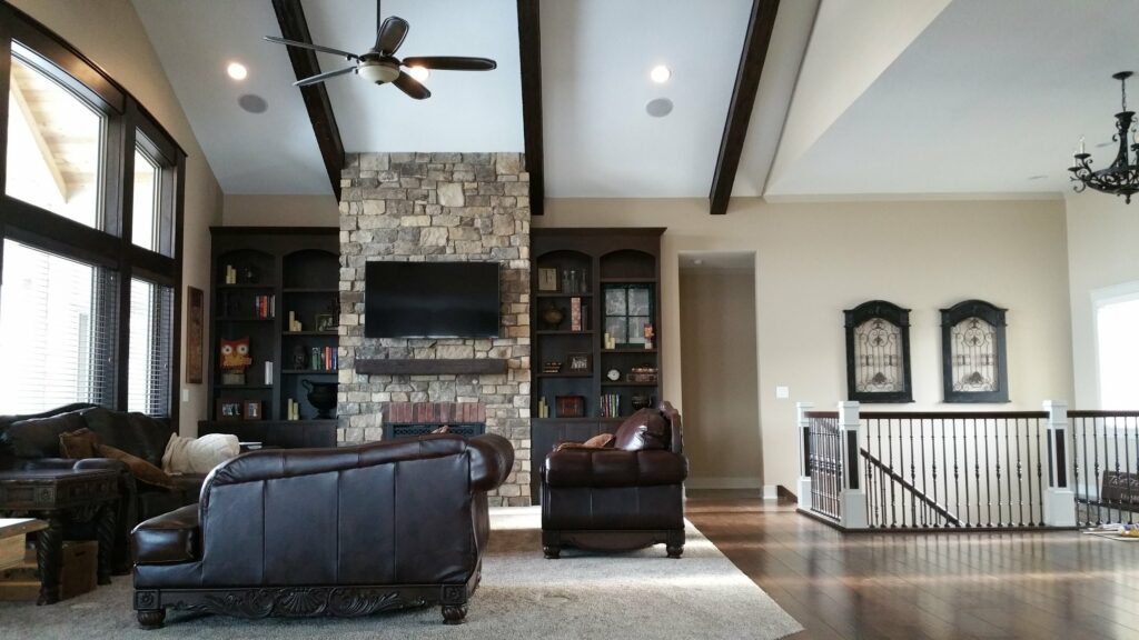 A spacious living room with vaulted ceilings, wooden beams, and large windows. It has a stone fireplace with a TV mounted above, flanked by built-in bookshelves. There are dark leather sofas, a light beige rug, and a staircase with a black railing in the background.
