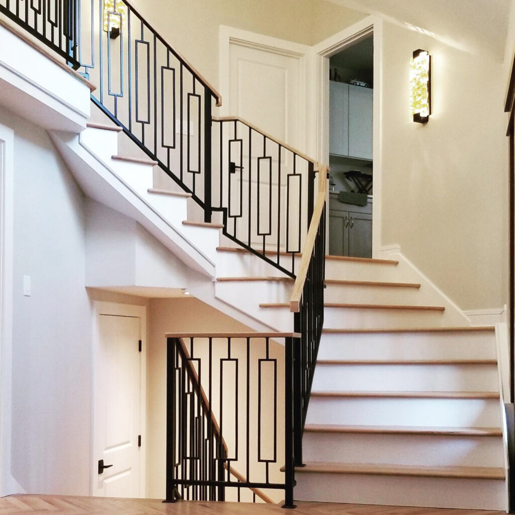 A modern staircase with black metal railings and wooden handrails. The stairs split into two directions, leading up to a landing with an open door revealing a kitchen area. The walls are painted light gray and a wall sconce light hangs on the right.