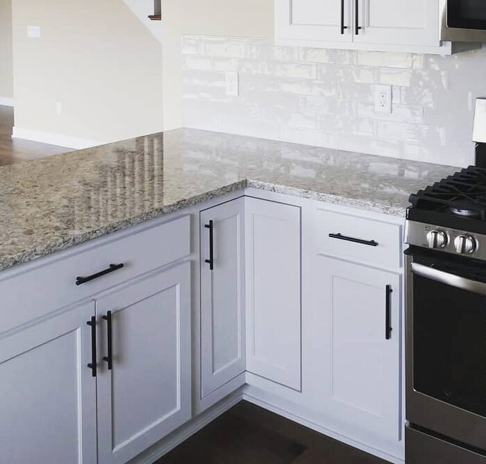 A modern kitchen with white cabinets, granite countertops, and stainless steel appliances, including a gas stove and microwave. A striped dish towel hangs on the oven handle. In the background, there's a staircase leading to an upper level and an open living area.