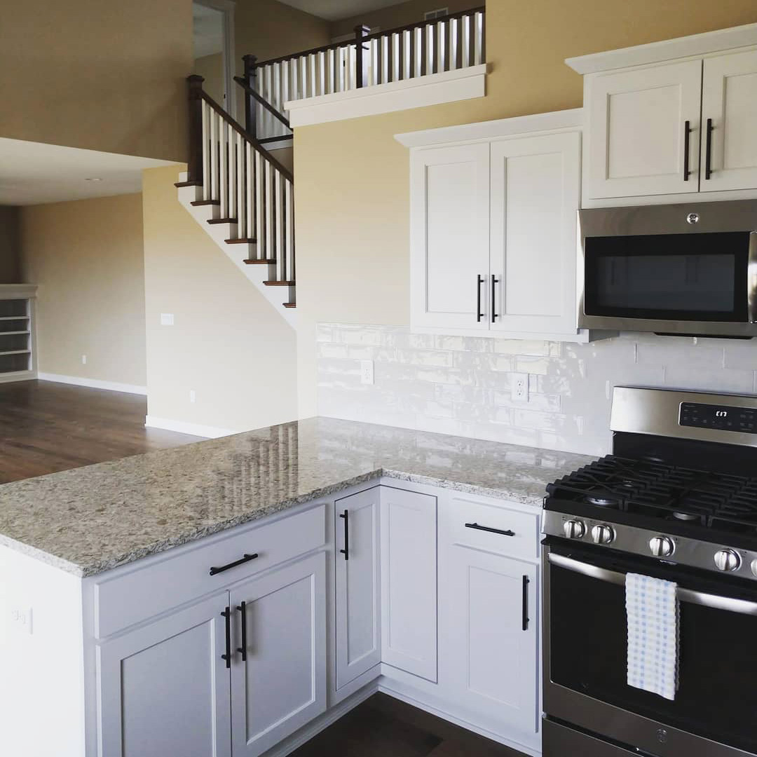 A modern kitchen with white cabinets, granite countertops, and stainless steel appliances, including a gas stove and microwave. A striped dish towel hangs on the oven handle. In the background, there's a staircase leading to an upper level and an open living area.