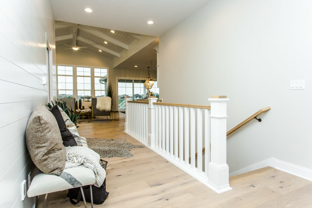 A modern, bright hallway with light wood floors and white walls. A staircase with a white railing descends to the right. To the left, a cushioned bench with pillows sits against the wall. The hallway leads to a spacious, open living area with large windows.
