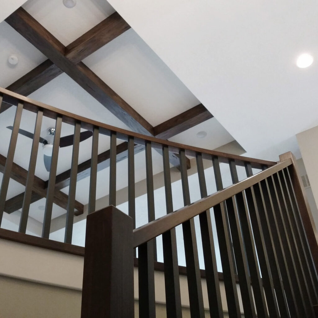 A modern interior features dark wooden ceiling beams, a ceiling fan, and recessed lighting. A dark wooden staircase with vertical slats in the foreground leads up to the upper level. The walls and ceiling are painted in neutral tones.