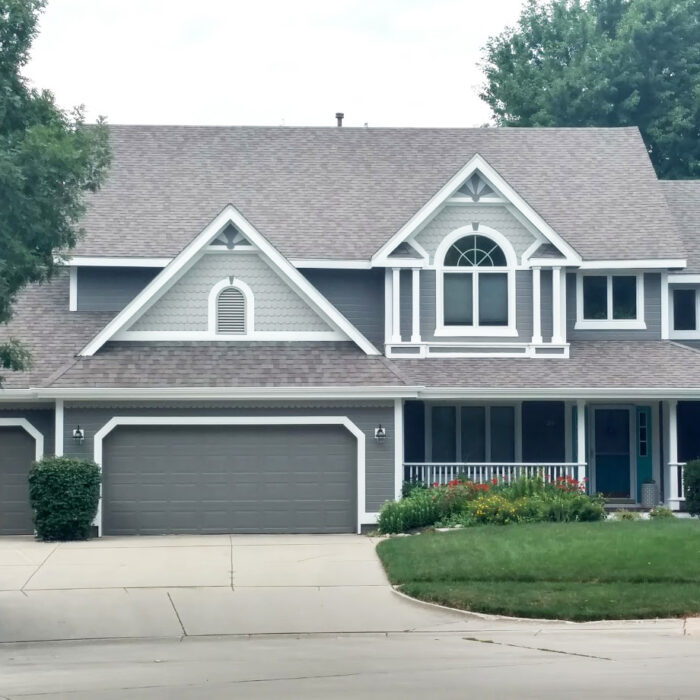 A suburban two-story house with gray siding, white trim, and a gable roof. It features a front porch, three-car garage with gray doors, and a well-maintained yard with bushes and green grass.