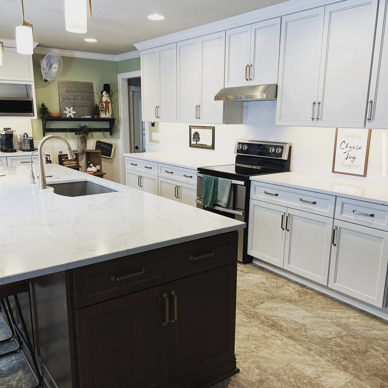 A modern kitchen with white cabinets, a stainless steel stove, and a marble countertop island with a sink in the center. A sign on the counter reads "Choose Joy." The backsplash is light-colored, and various decor items are displayed on the walls and counters.