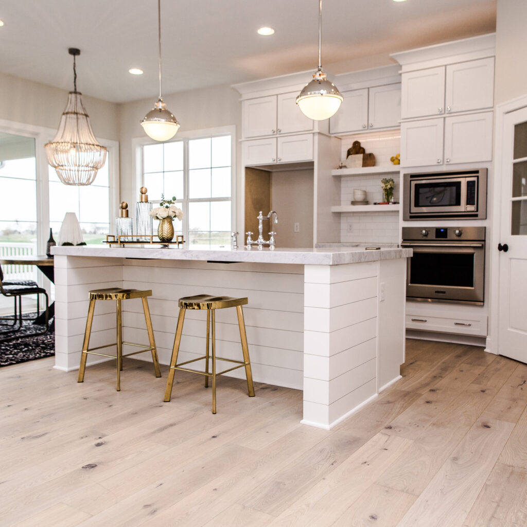 Modern kitchen with white cabinets, light wood flooring, and stainless steel appliances. Two gold-toned bar stools are positioned at a shiplap island with hanging pendant lights above. A large window provides natural light, and a decorative chandelier hangs nearby.
