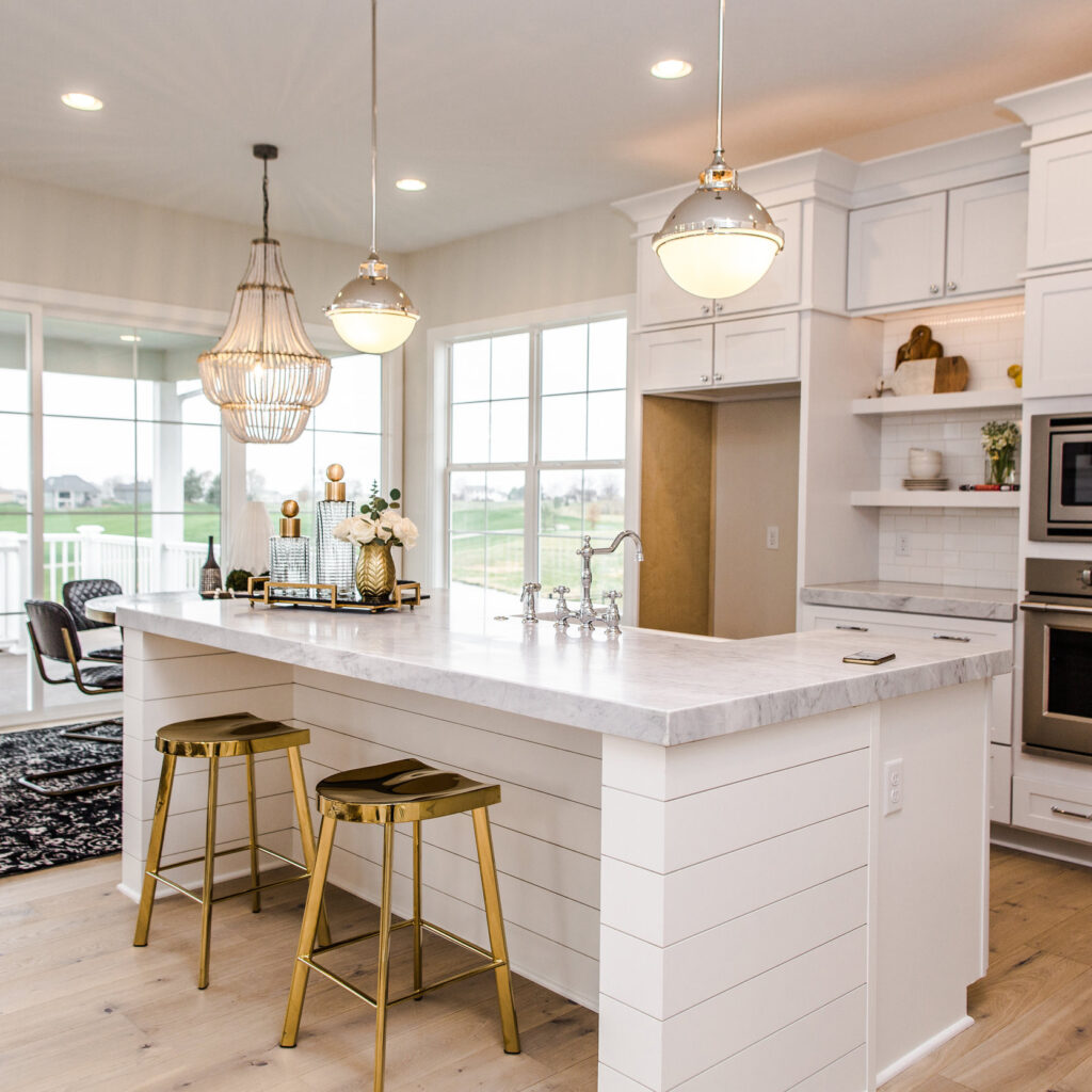 A modern kitchen features a large marble island with two gold barstools. Hanging pendant lights and a chandelier illuminate the space. There are white cabinets, open shelves, a built-in oven, and a dining area with large windows in the background.