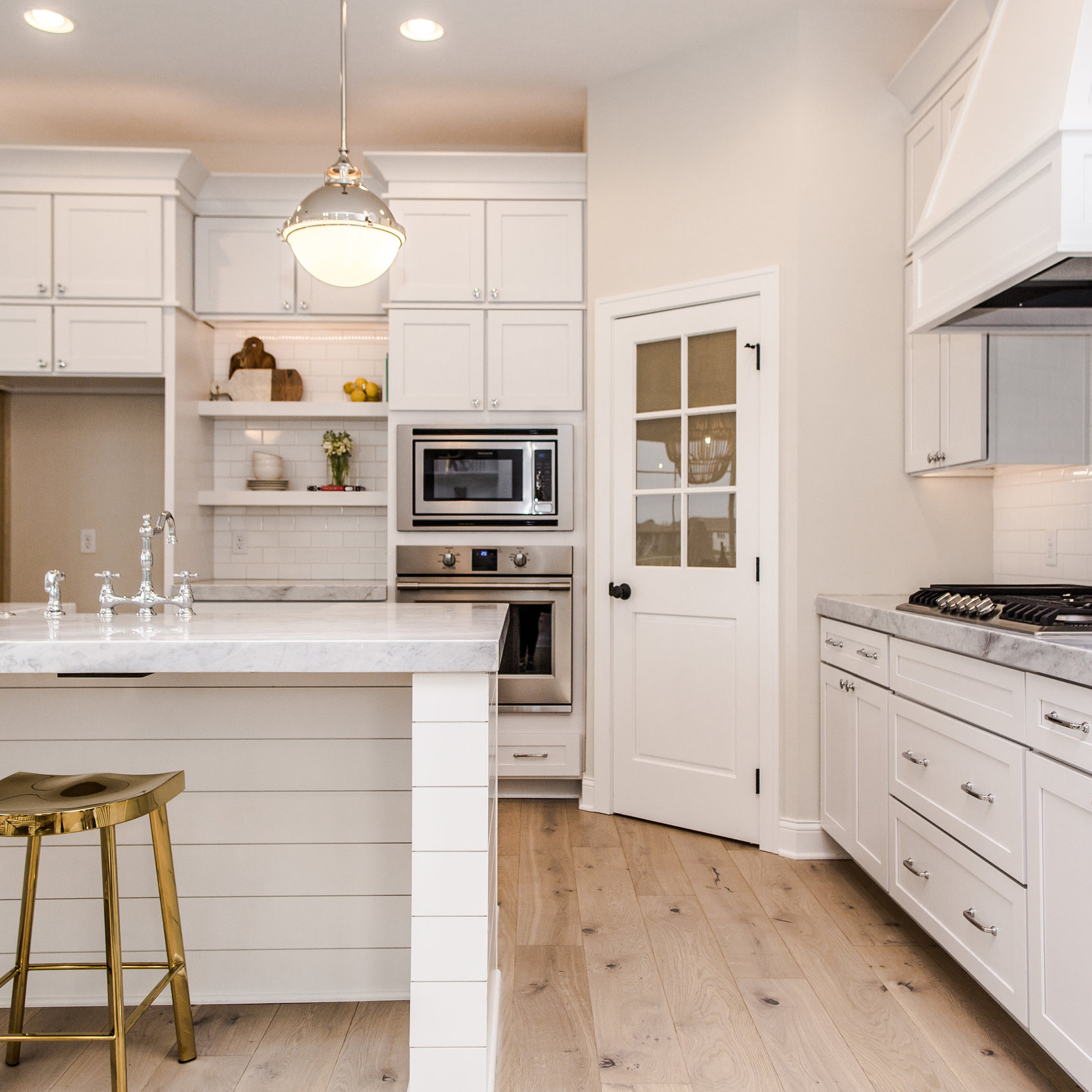 A modern kitchen with white cabinetry, a marble countertop island, and stainless steel appliances. The island has a golden stool, and a pendant light hangs above it. The kitchen features light wood flooring and a door with a frosted glass panel.