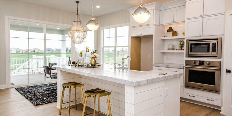 A modern kitchen with white cabinets and a large marble island equipped with two gold stools. Pendant lights hang above the island, and there are stainless steel appliances built into the cabinetry. A large window and glass door offer a view of a patio and green yard.