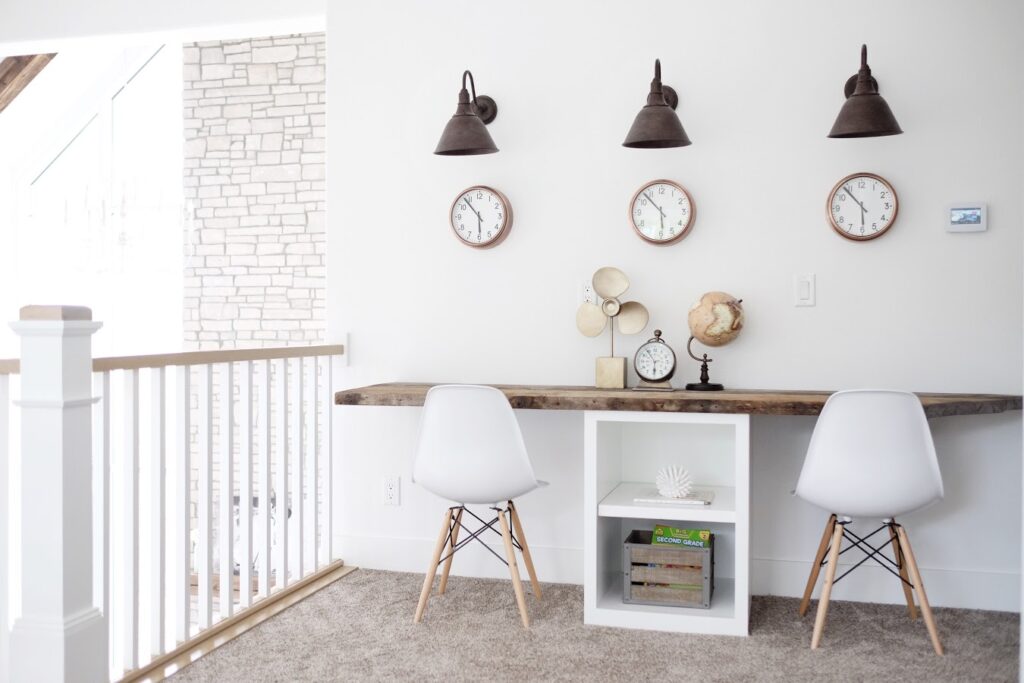 A minimalist workspace features a wooden desk with storage shelves and two white modern chairs. The wall above the desk is adorned with three clocks and industrial-style light fixtures. The area is bright and airy, with a railing and a partially visible staircase.