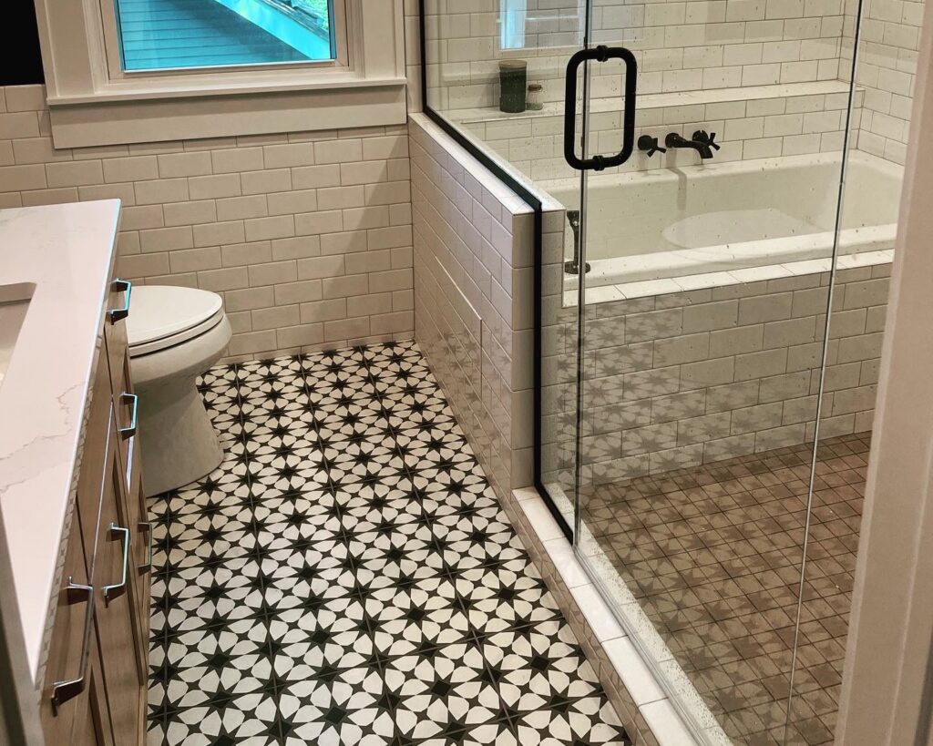 A modern bathroom with a glass shower enclosure and a bathtub inside it, white subway tile walls, a window with a view of the green outside, a black and white patterned tile floor, a toilet, and a white countertop with cabinets beneath it.