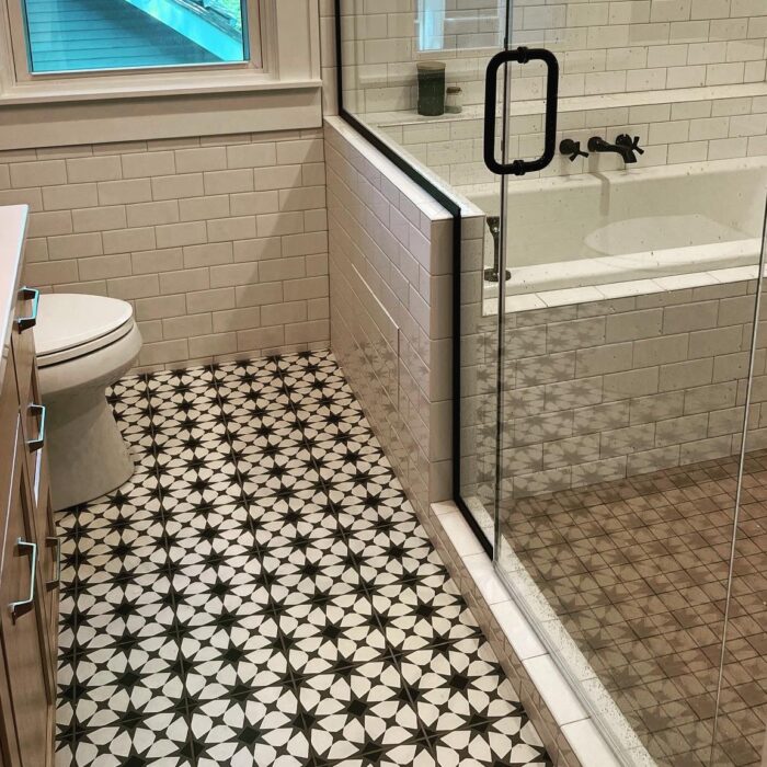 A modern bathroom with a glass shower enclosure and a bathtub inside it, white subway tile walls, a window with a view of the green outside, a black and white patterned tile floor, a toilet, and a white countertop with cabinets beneath it.