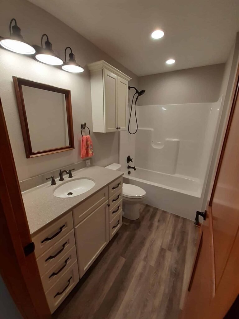 A modern bathroom with a wooden floor, white cabinet and drawers under a countertop sink, a mirror with a wooden frame, a toilet, and a white bathtub with a curved shower rod and black fixtures. Three lights are mounted above the mirror.