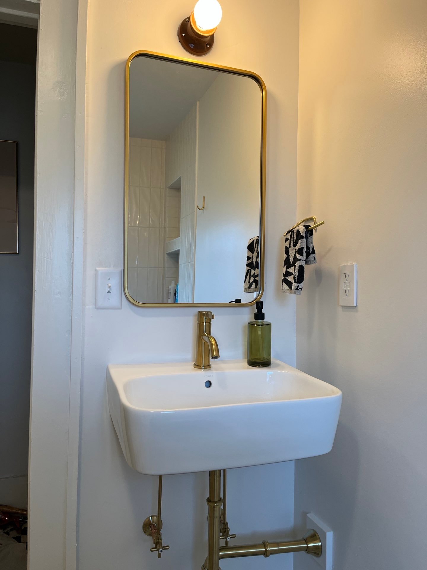 A modern bathroom sink with a gold faucet and a rectangular mirror with a gold frame. A small hand towel with a black and white pattern hangs on a hook to the right of the mirror. A green liquid soap dispenser rests on the sink. A single bulb light fixture is above the mirror.