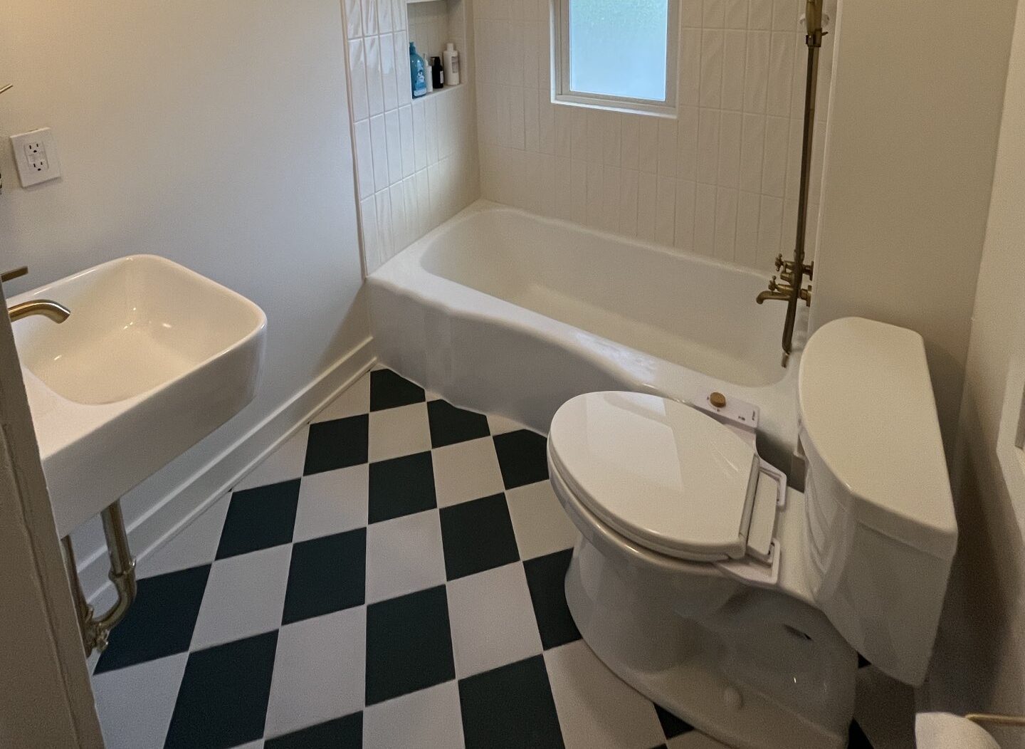 A small bathroom with a black-and-white checkered floor. It features a white bathtub with a wall-mounted shower alongside a small white sink with a golden tap and a white toilet. The window above the bathtub provides natural light, and built-in shelves hold toiletries.