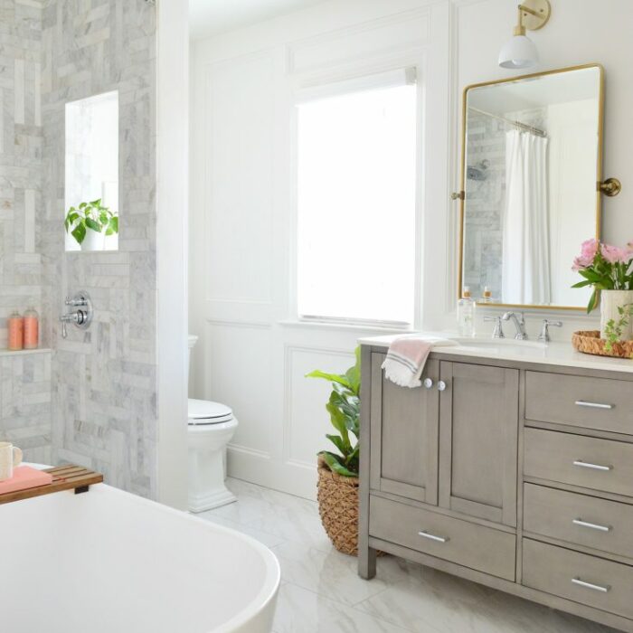A bright bathroom with a freestanding bathtub in the foreground. On the left, there's a shower with gray and white marble tiling. A toilet is near a window with sheer curtains. A vanity with a large mirror, potted plant, and flowers sits by the sink on the right.