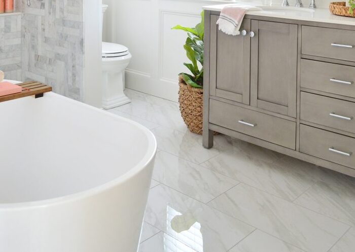 A bright bathroom with a freestanding bathtub in the foreground. On the left, there's a shower with gray and white marble tiling. A toilet is near a window with sheer curtains. A vanity with a large mirror, potted plant, and flowers sits by the sink on the right.