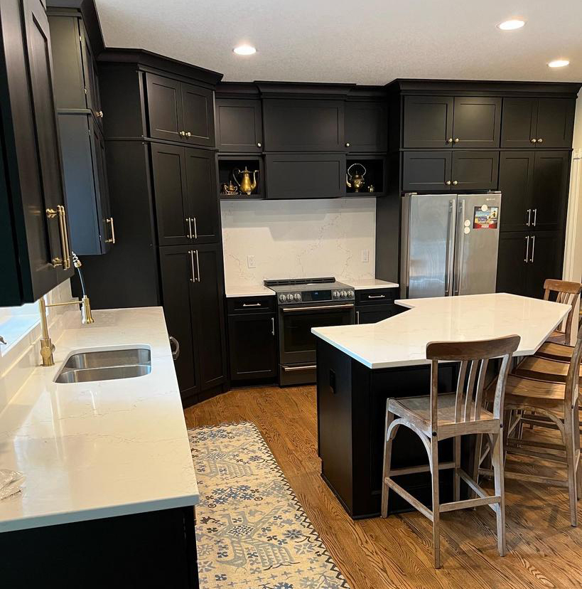 A modern kitchen featuring dark cabinets, a stainless steel refrigerator, a range oven, and white countertops. A wooden island with bar stools and a patterned rug add warmth to the space. Recessed lighting illuminates the area.