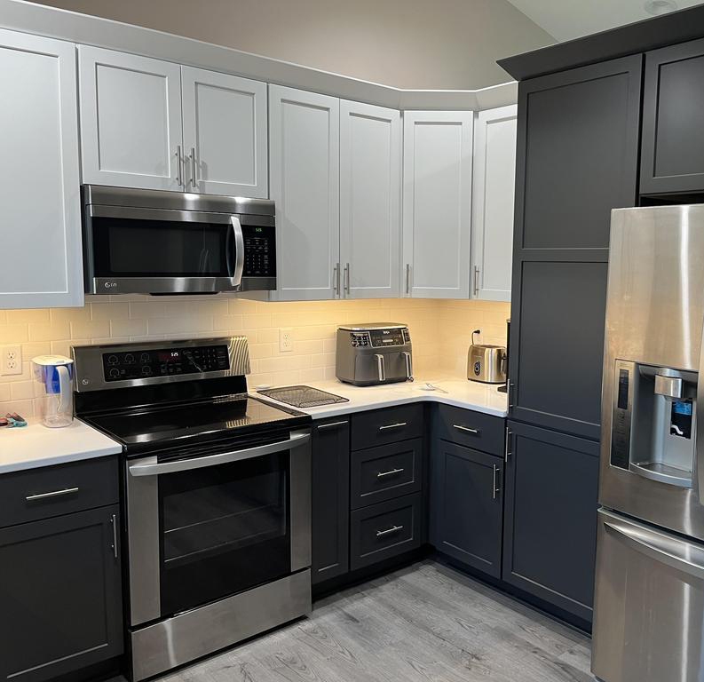 A modern kitchen with a stainless steel refrigerator, stove, microwave, and toaster oven. The cabinetry is two-toned, with white upper cabinets and dark lower cabinets. The countertops are white, and the backsplash is a light-colored tile. The floor is a light wood laminate.