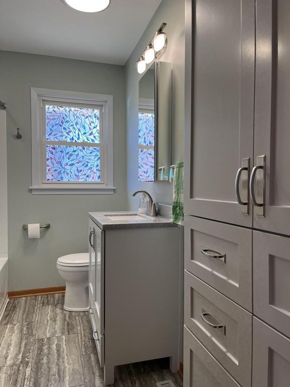 A modern bathroom features grey tiled flooring, white cabinetry, and a matching vanity with a sink and mirror. A toilet is positioned by a window with frosted, patterned glass. Overhead lighting and a green bottle of hand soap add to the clean, bright look.