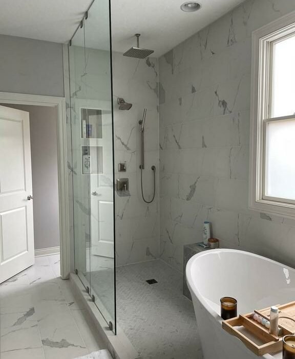 A modern bathroom features a glass-enclosed shower with a rainfall showerhead, grey and white marble walls, and a freestanding white bathtub. A wooden tray rests on the tub, holding a book and a candle. A window on the right lets in natural light.