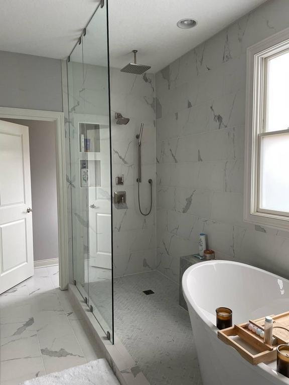 A modern bathroom features a glass-enclosed shower with a rainfall showerhead, grey and white marble walls, and a freestanding white bathtub. A wooden tray rests on the tub, holding a book and a candle. A window on the right lets in natural light.