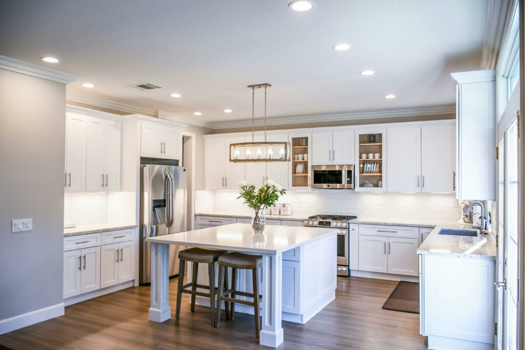 A Reese Builders modern kitchen with white cabinets, stainless steel appliances, and a central island. The island has two wooden stools and a vase with flowers. Recessed lighting and a rectangular chandelier illuminate the space. Light wood flooring complements the white decor.