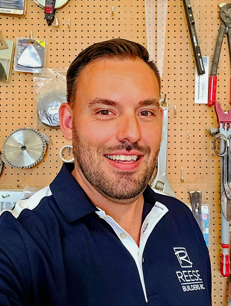 Smiling man in a dark blue polo shirt with a company logo stands in a workshop. Tools like saw blades, wrenches, and measuring tapes are organized on a pegboard behind him.