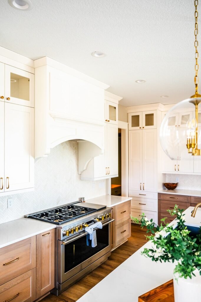 Bright, modern kitchen with white and wood cabinets, stainless steel stove, herringbone backsplash, gold hardware, a hanging glass light fixture, and a potted plant on a white countertop.