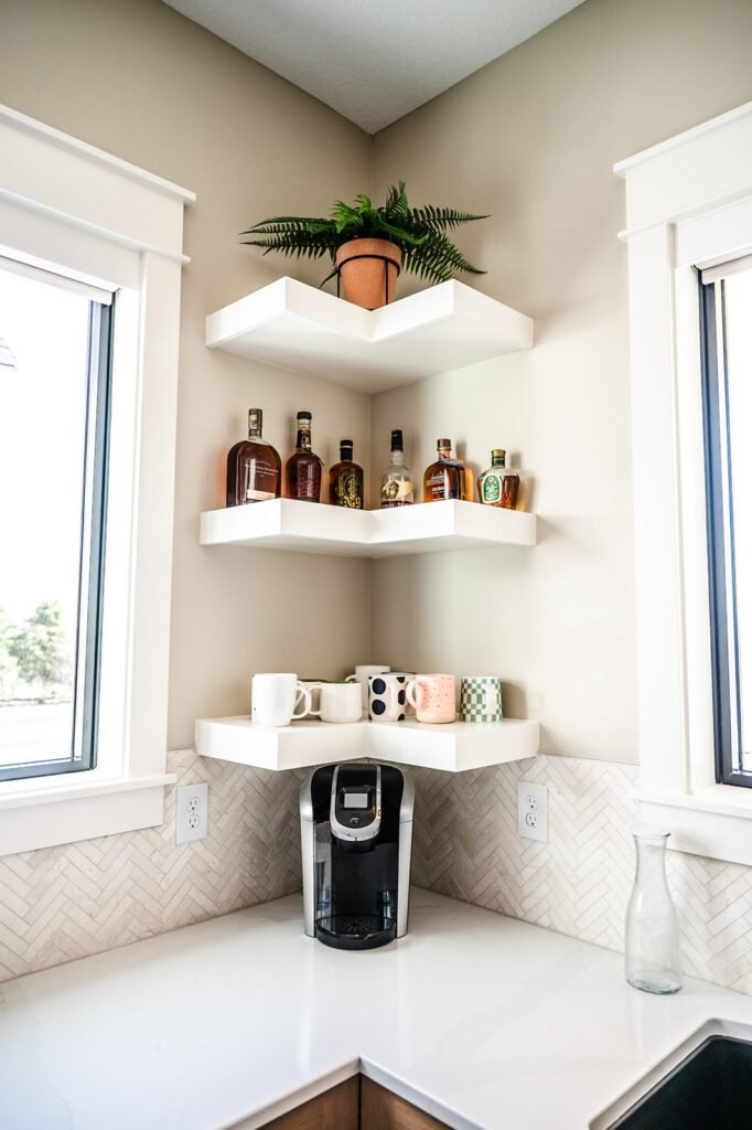 A corner kitchen countertop with a coffee maker, four patterned mugs, and floating shelves above holding bottles of liquor and a potted fern. Sunlight comes through two windows flanking the corner.