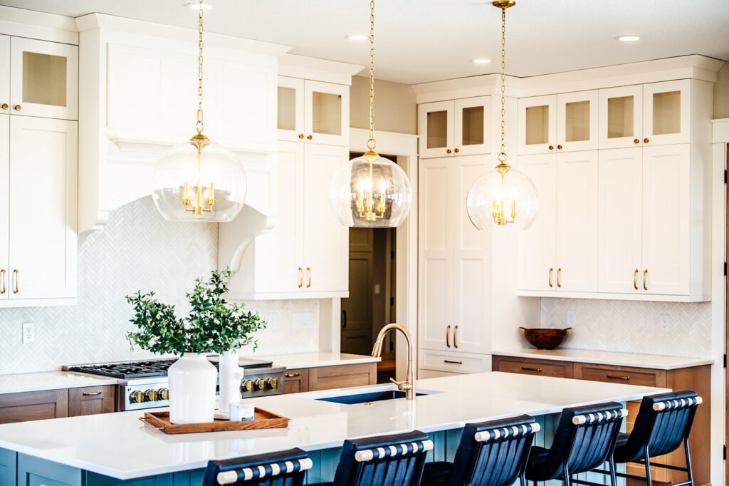 Bright, modern kitchen with white cabinets, gold hardware, a large island topped with a tray holding vases, three glass pendant lights, and black barstools lined up at the counter.