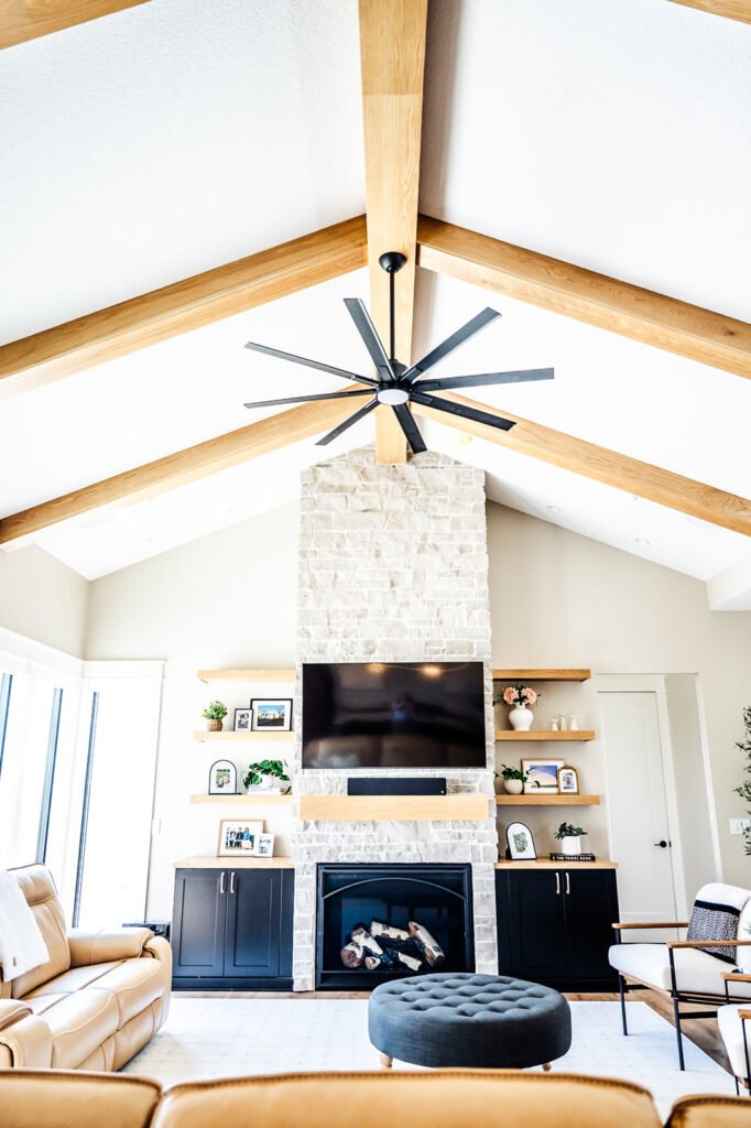 Bright living room with high ceilings, exposed wooden beams, a large ceiling fan, a stone fireplace with a TV mounted above, built-in cabinets and shelves, tan leather sofas, and large windows letting in natural light.