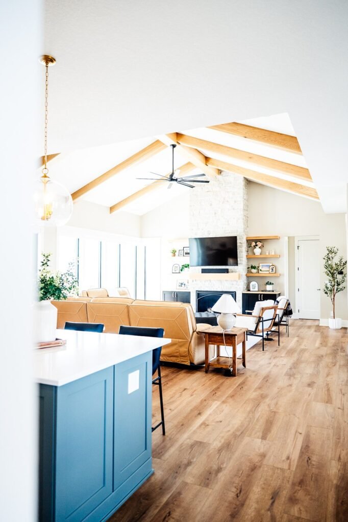 Bright, modern living room with vaulted ceiling, exposed wood beams, light wood flooring, a blue kitchen island, tan sofa, wall-mounted TV above a stone fireplace, large windows, and minimalist decor. Des Moines Home Remodel