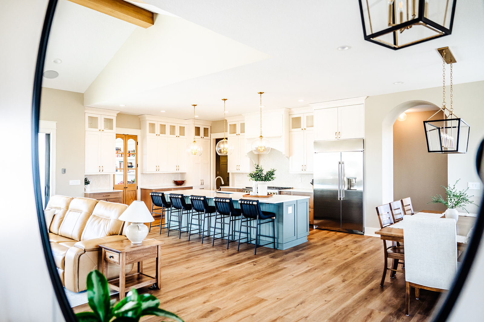 Bright, modern open-concept kitchen and living area with white cabinets, stainless steel appliances, a blue island with black chairs, tan sofa, wooden dining table, and large windows letting in natural light.