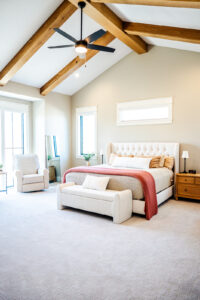 Bright, spacious bedroom with a large bed, white upholstered headboard, coral throw blanket, and a small bench. Wooden ceiling beams and neutral decor create a cozy, modern atmosphere. A chair and nightstands complete the space. Reese Builders, Luxury Bedroom, Wood Beams, Des Moines, Iowa