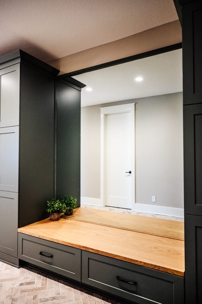 A modern entryway with dark cabinets, a wooden bench, two small potted plants, and a large wall mirror reflecting a white door and beige walls. The floor has a herringbone tile pattern.