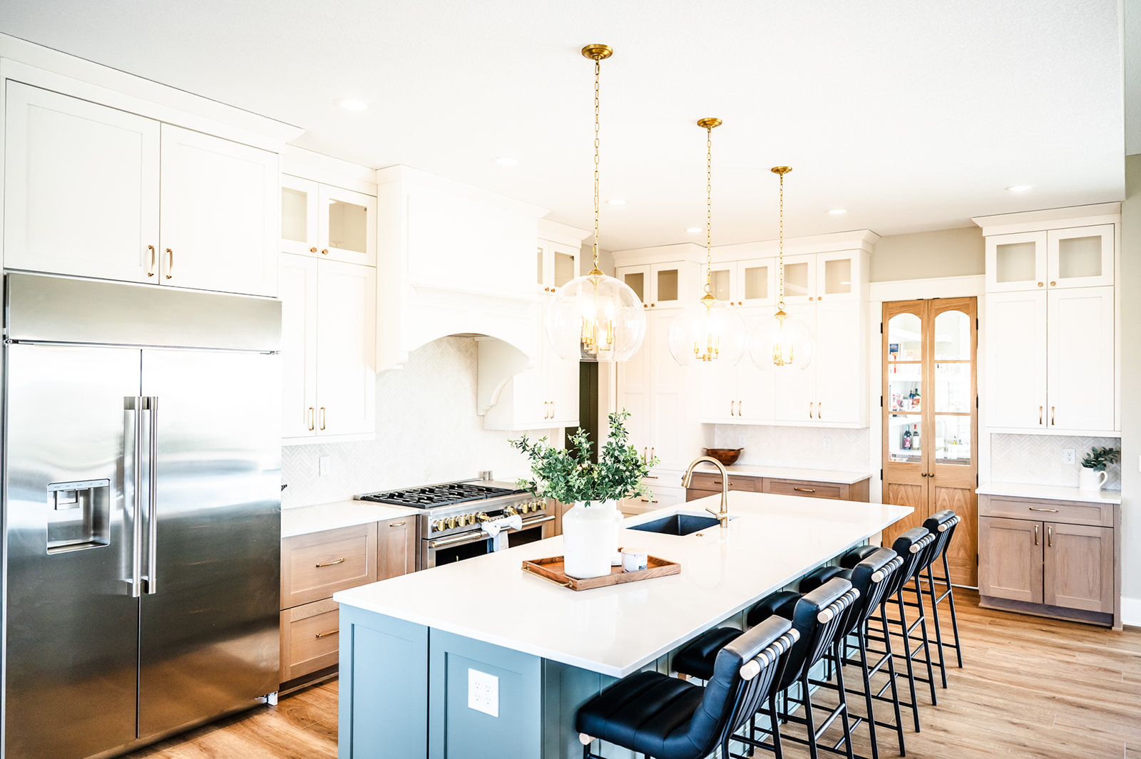 Bright modern kitchen with white cabinets, stainless steel appliances, a large island with a white countertop, black barstools, three pendant lights, and wooden accents. Greenery decorates the island.