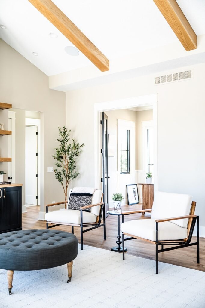 Bright, modern living room with white walls, wooden ceiling beams, two white armchairs with black frames, a round dark ottoman, a side table, potted plants, and large windows letting in natural light.