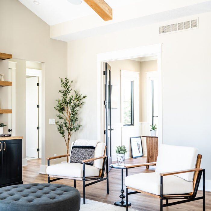 Bright, modern living room with white walls, wooden ceiling beams, two white armchairs with black frames, a round dark ottoman, a side table, potted plants, and large windows letting in natural light.