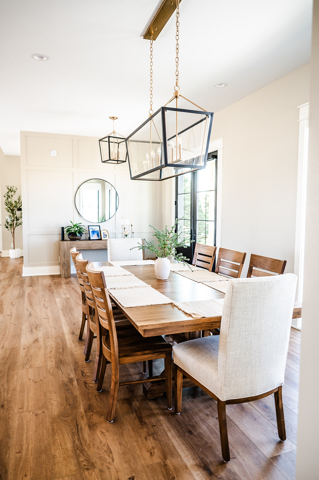 Des moines remodel, reese builders, kitchen, Modern dining room with a wooden table, eight chairs, two geometric pendant lights, and a white vase centerpiece. Light wood floors, beige walls, and a mirror with plants in the background create a bright, airy space.