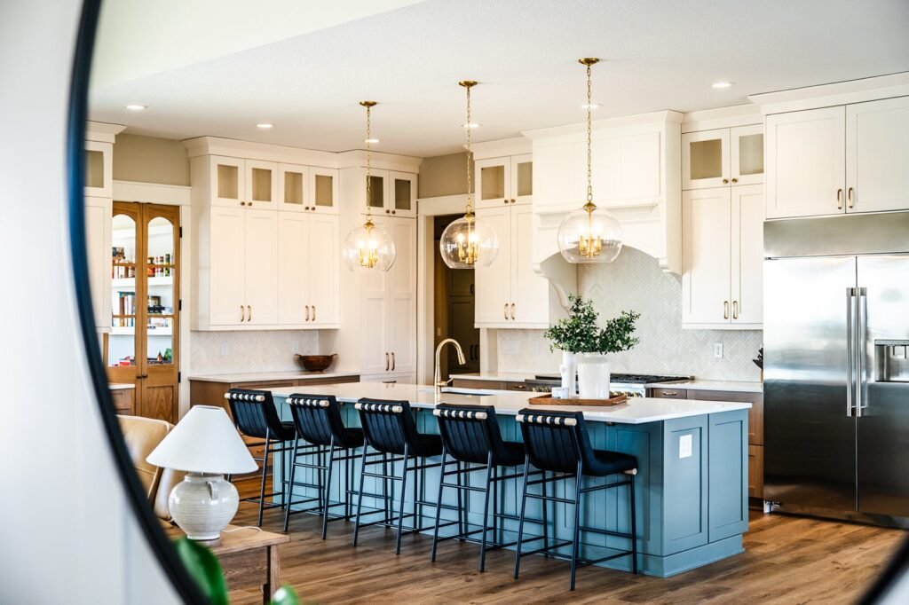 Modern kitchen with white cabinets, a large blue island with bar stools, pendant lights, stainless steel appliances, and wooden floors. A plant and decorative items are on the island, and shelves are visible in the background.