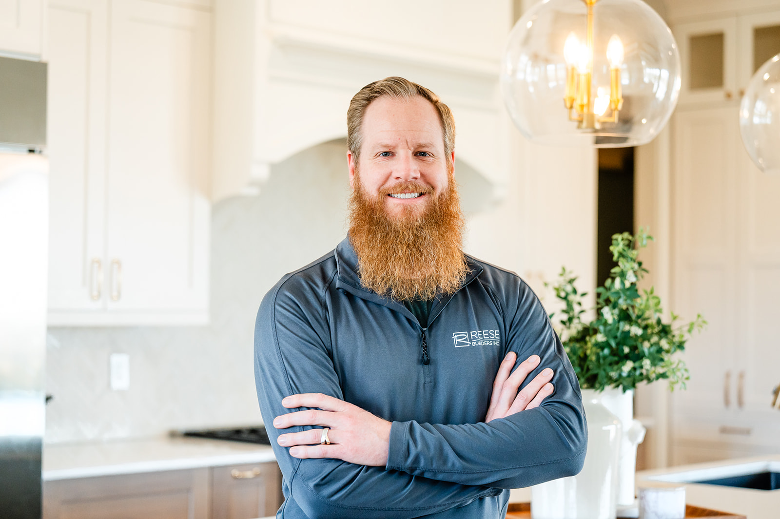 A bearded man with reddish hair stands smiling with arms crossed in a bright kitchen. He wears a gray zip-up top with a “Reese” logo. A modern light fixture and a vase with greenery are visible in the background.
