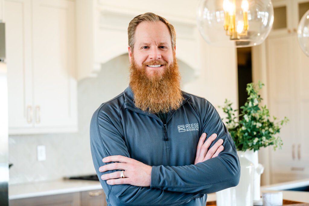 A man with a long red beard stands smiling in a modern kitchen, arms crossed, wearing a gray zip-up shirt with a "Reese" logo. Bright lighting and green plants decorate the background.