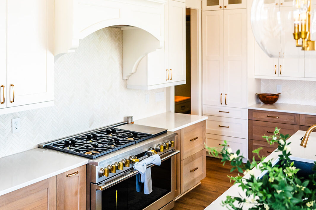 Bright, modern kitchen with white and wood cabinets, stainless steel stove, herringbone backsplash, gold hardware, a hanging glass light fixture, and a potted plant on a white countertop.