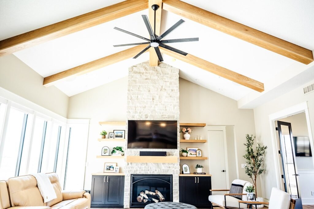Bright living room with vaulted ceiling, exposed wooden beams, a large black ceiling fan, a stone fireplace, wall-mounted TV, built-in shelves with décor, and modern furniture. Large windows let in natural light in remodeled home in Huxley Iowa